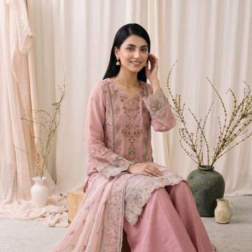 Woman in a pink traditional outfit sitting on a light-colored floor with decorative plants and curtains in the background.